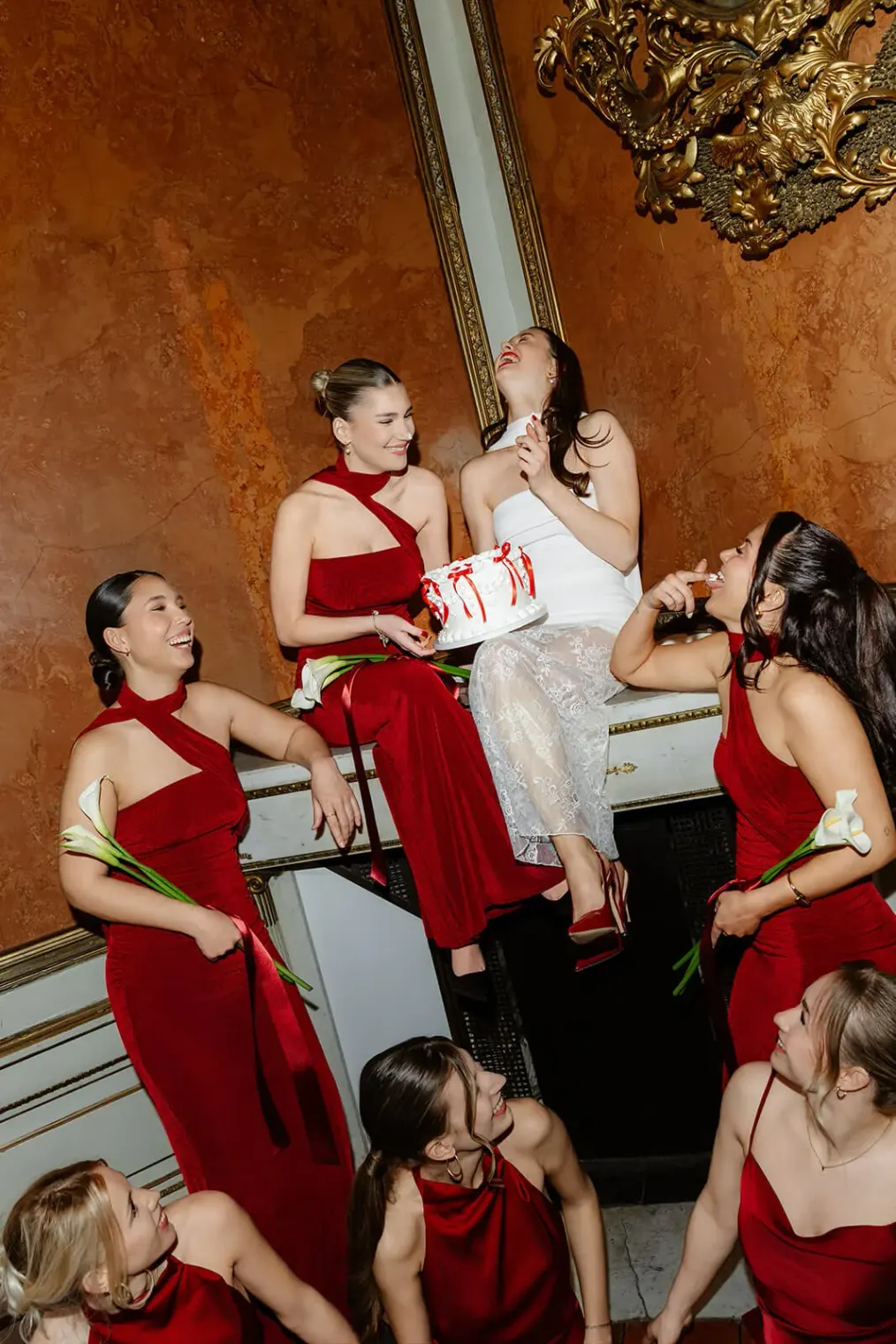 Braut sitzt lachend mit Brautjungfern in roten Kleidern und Hochzeitstorte